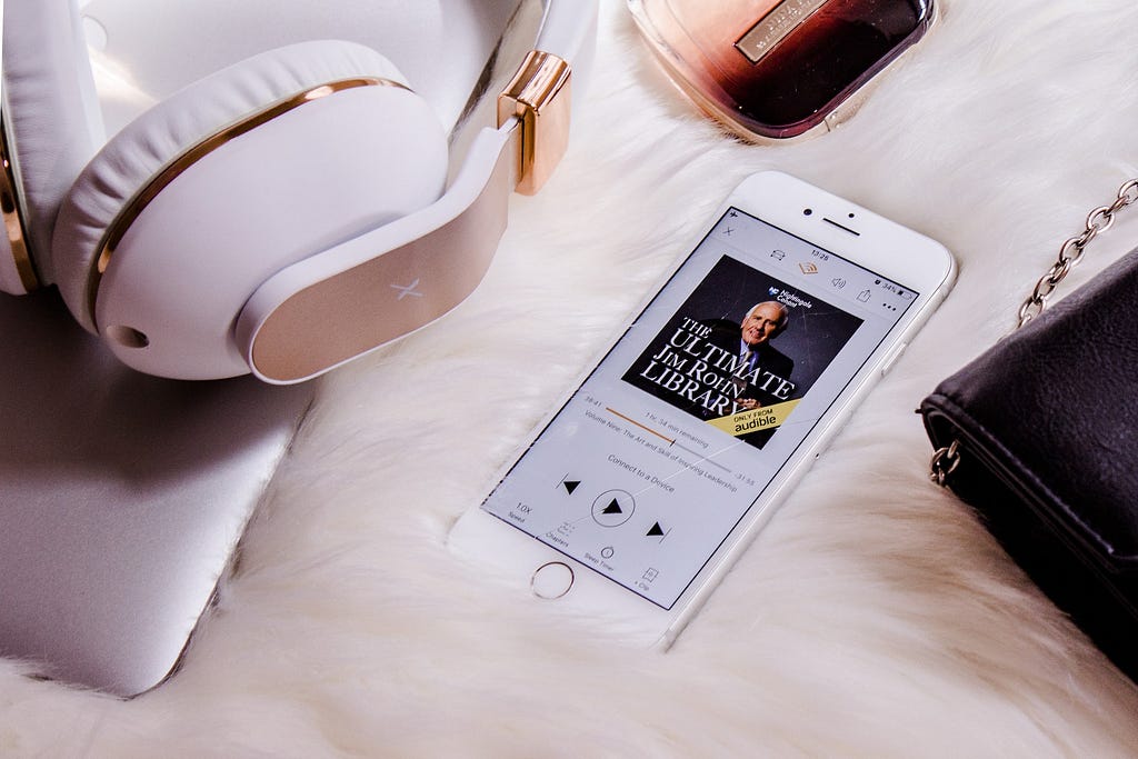 A iPhone with an audiobook visible on the screen, lying on a cozy white background, headphones, a bag and other stuff nearby.