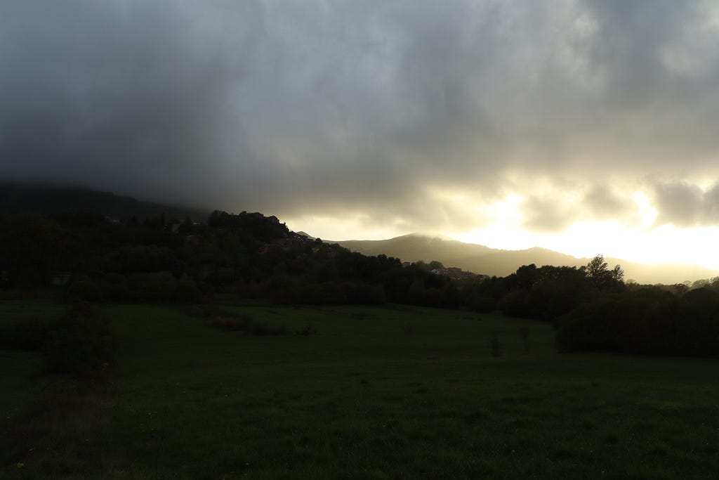 A moody twilight scene of rolling green hills and a small village silhouette beneath heavy, dark clouds, with a faint golden light breaking through in the distance.