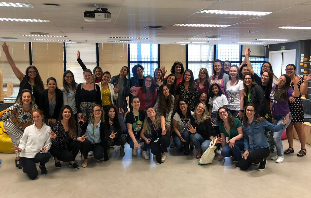 Foto de 37 mulheres colaboradoras do Sicredi, todas sorrindo, com as mãos e braços para cima em uma pose animada e em um ambiente empresarial.
