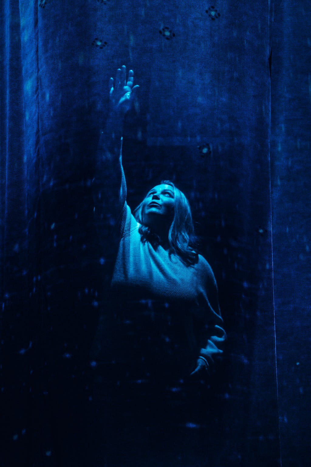 Amy Rietsma, a white actor in a light colored sweater, is reaching upwards with her left hand from behind a transparent curtain. The whole photo is bathed in blue, like light shining through to the bottom of the sea.