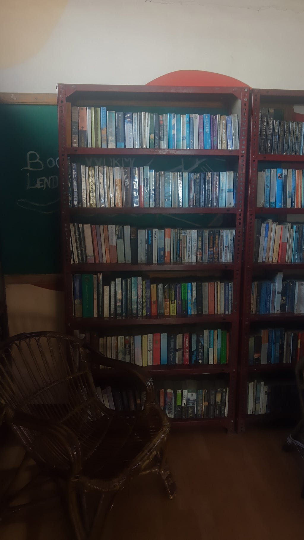 Tall shelves filled with books next to an empty wicker chair. The bookshelf is filled with various books and the chair positioned in front of it to the left.