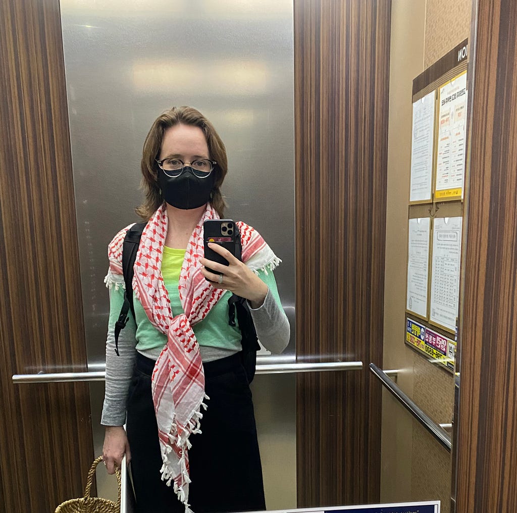The author, a white middle aged woman with medium brown hair takes a selfie in an elevator mirror. She is wearing a colorful shirt and a red and white Keffiyeh with a black skirt, black backpack, and black mask.