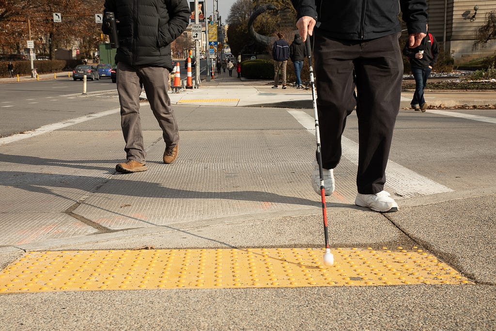 Two people crossing an intersection with the image taken from a low angle showing the curb ramp. One person uses an assistive can typically used by people with visual impairments.