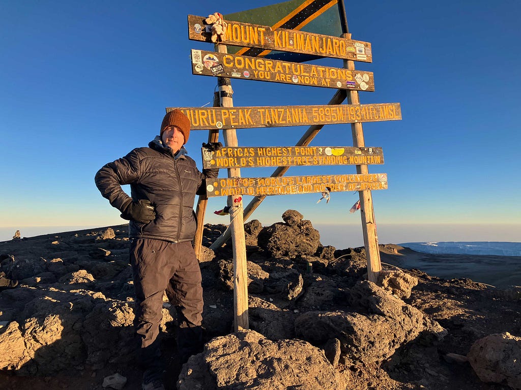 Author standing next to the summit sign at the top of Mount Kilimanjaro at sunrise under deep blue sky.