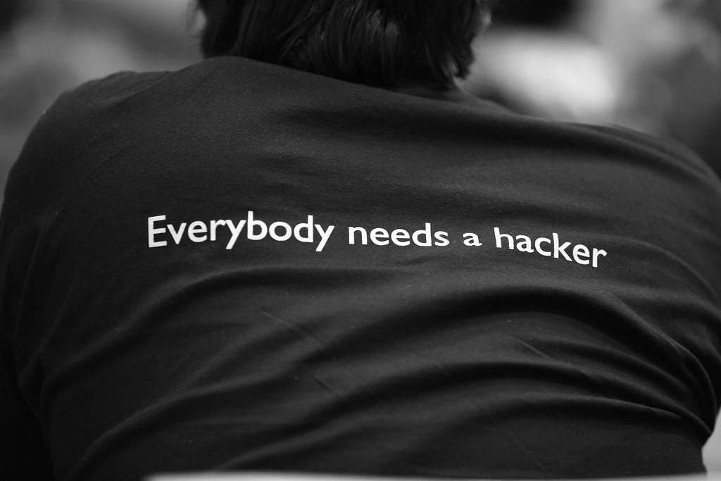 Black and white image of a person wearing a black t-shirt that says “Everybody needs a hacker”