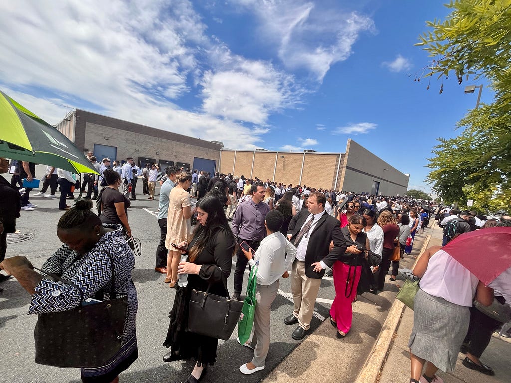 Color photo of a bright sunny blue skies day where tons of people of varied adult ages, sizes, gender, culture, and attire, with some taking cover under personal umbrellas are shown in long lines outside a big building.