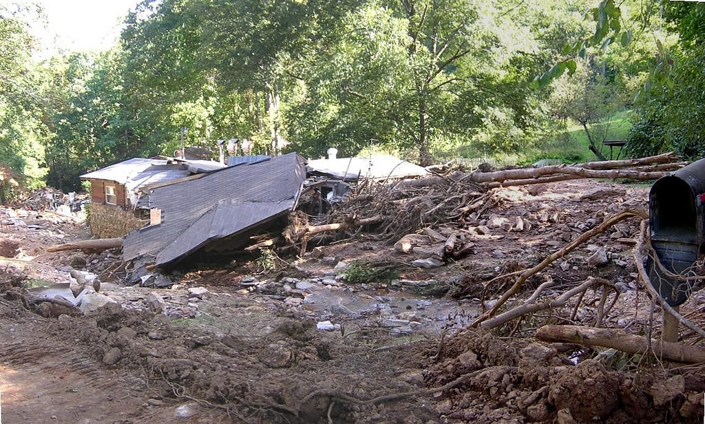 A house with its roof lying detached and boulders, trees, and other debris strewn across the foreground