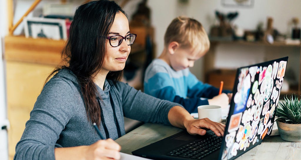 A woman working at home with her child playing beside her.