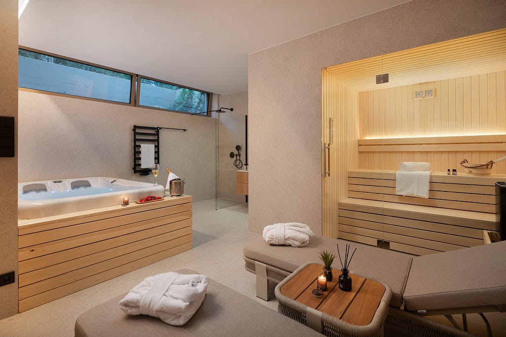 A luxurious spa room featuring a modern design with light wood accents. On one side, a built-in hot tub with clean lines and a wooden exterior is filled with bubbling water. Next to it, a glass-enclosed sauna with sleek wooden benches is illuminated by soft, warm lighting. In the foreground, there are comfortable loungers with plush cushions and fluffy white robes neatly placed on them. A small wooden table holds a candle, aromatic diffusers, and a plant, adding to the tranquil ambiance.