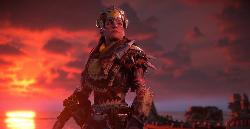 Image of Aloy from slightly below, showing her looking off to the distance with a gorgeous, orange sunset in the background. She is wearing yellow and blue Tenakth armor and has white Utaru facepaint.