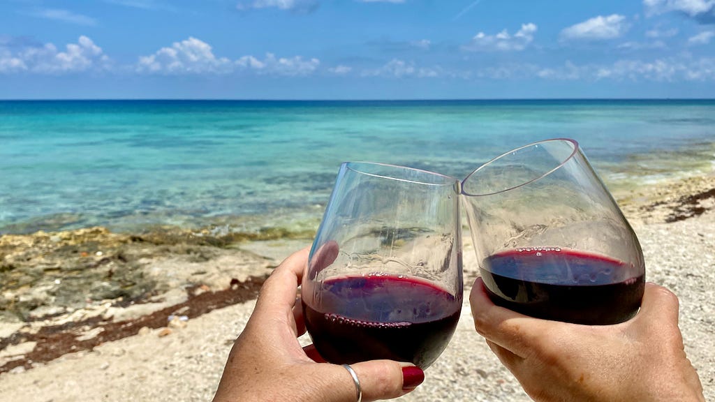 Cheers! Two stemless wine glasses with red wine clinking under the sun with gorgeous light turquoise Caribbean water in the background.