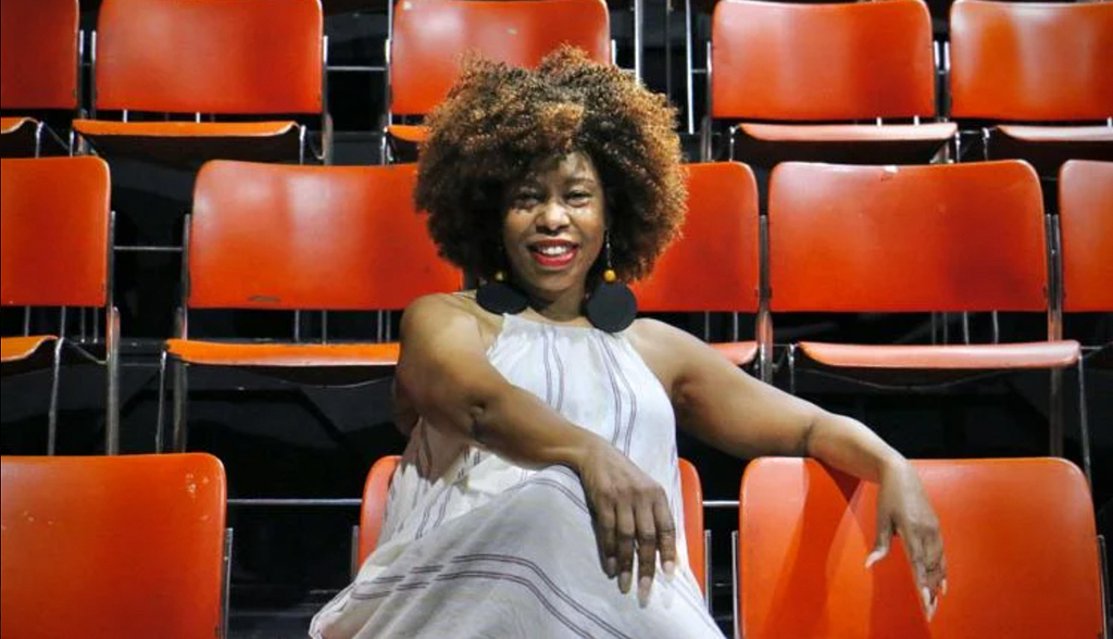 A woman with curly brown hair and brown skin in a white dress sits in a row of orange chairs and smiles at the camera.