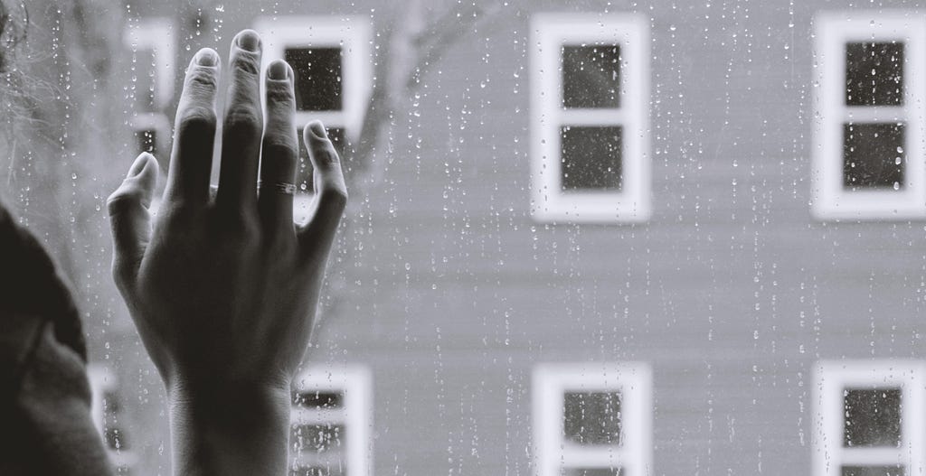 Black and white filter of a hand pressed against a window with raindrops on it.