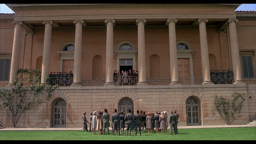A shot from Salo which shows the side of an enormous palace that fills the whole frame. On a balcony, the libertines and sex workers are gathered, talking to the captives and soldiers on the lawn below.