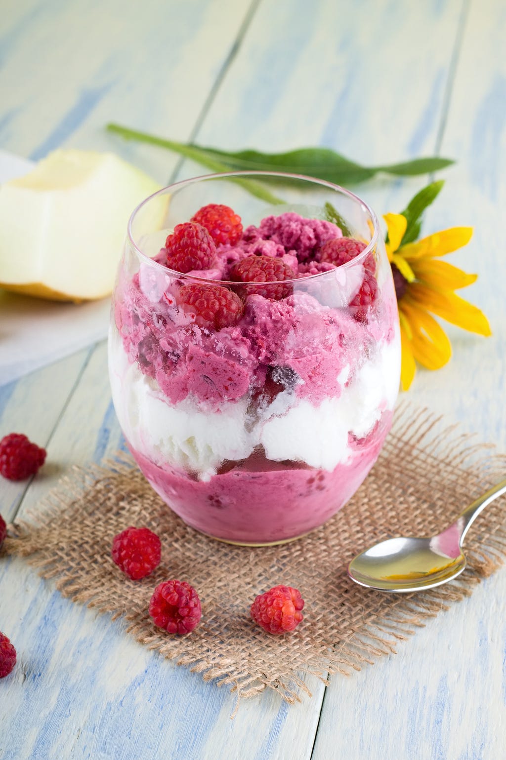 A cup of kindness — beautiful, brightly lit glass of white and raspberry colored ice cream, topped with raspberries and with a daisy laid nearby.