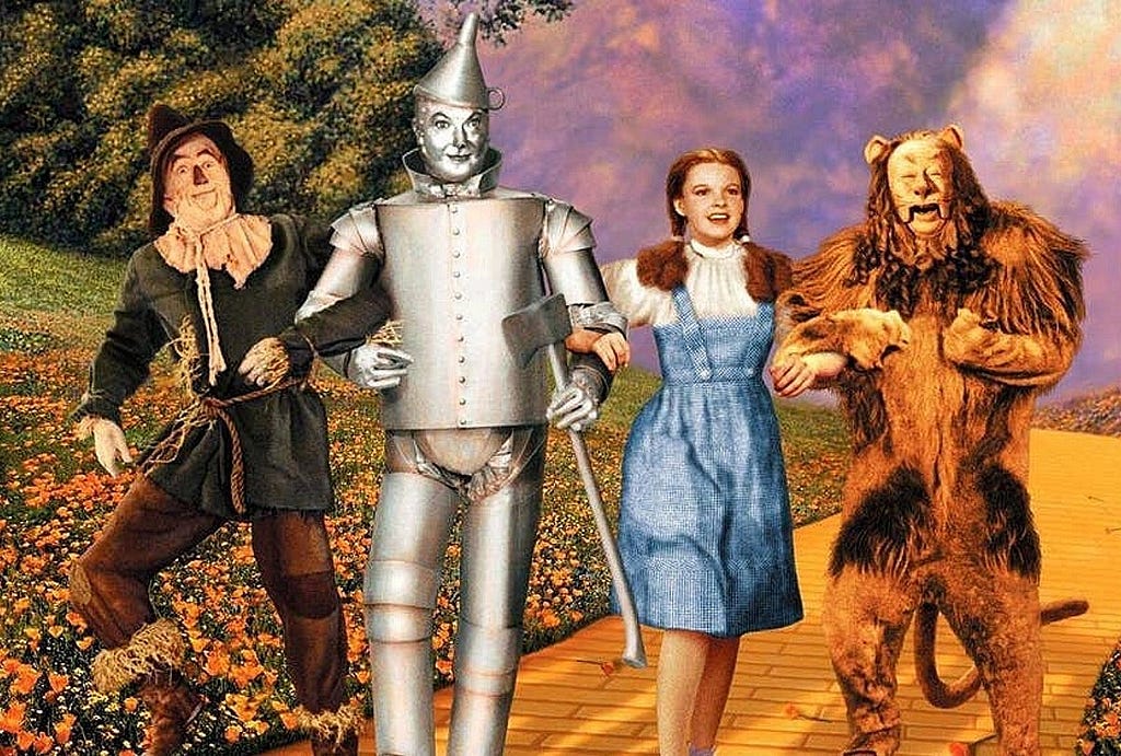 Movie still from The Wizard of Oz with the scarecrow, tin man, Dorothy, and the lion on the yellow brick road