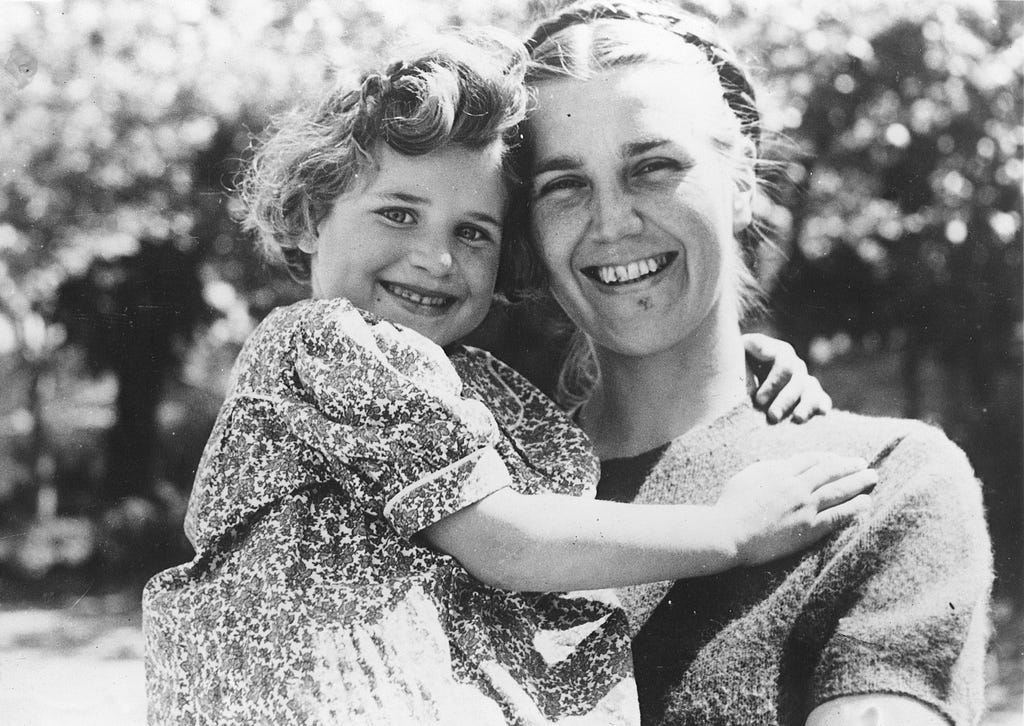 On a sunny day, a woman smiles while holding a smiling young girl in her arms. The girl smiles toward the camera and has her arm around the woman’s neck. She is wearing a flowered dress with puffed sleeves.