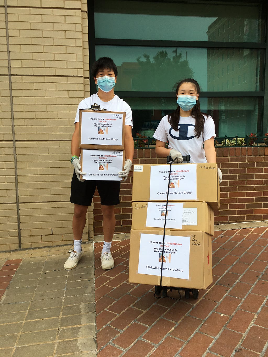 Two young people stand next to each other with masks on, holding boxes filled with kits.