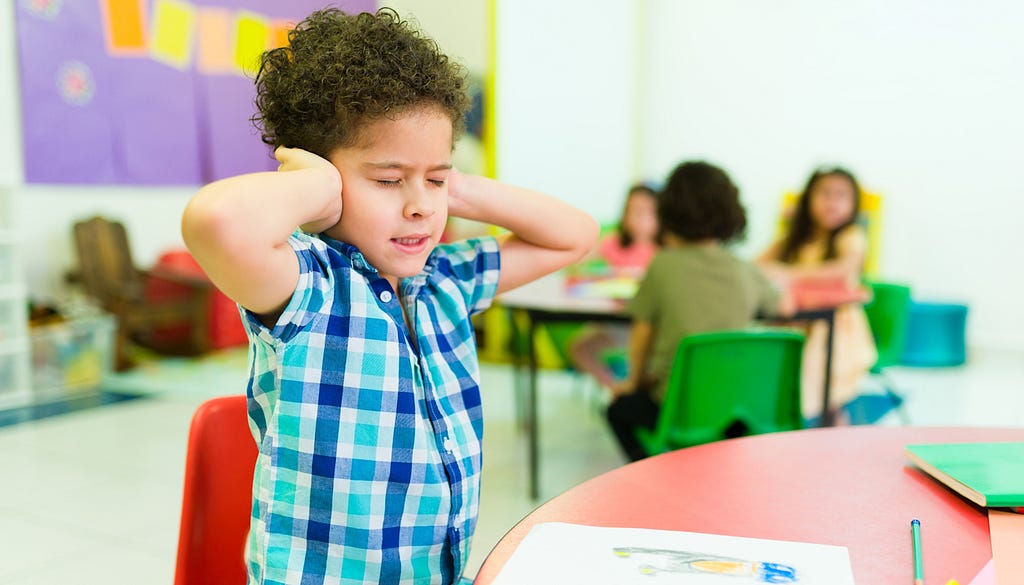 Autistic boy covers ears to block loud noise in preschool setting.