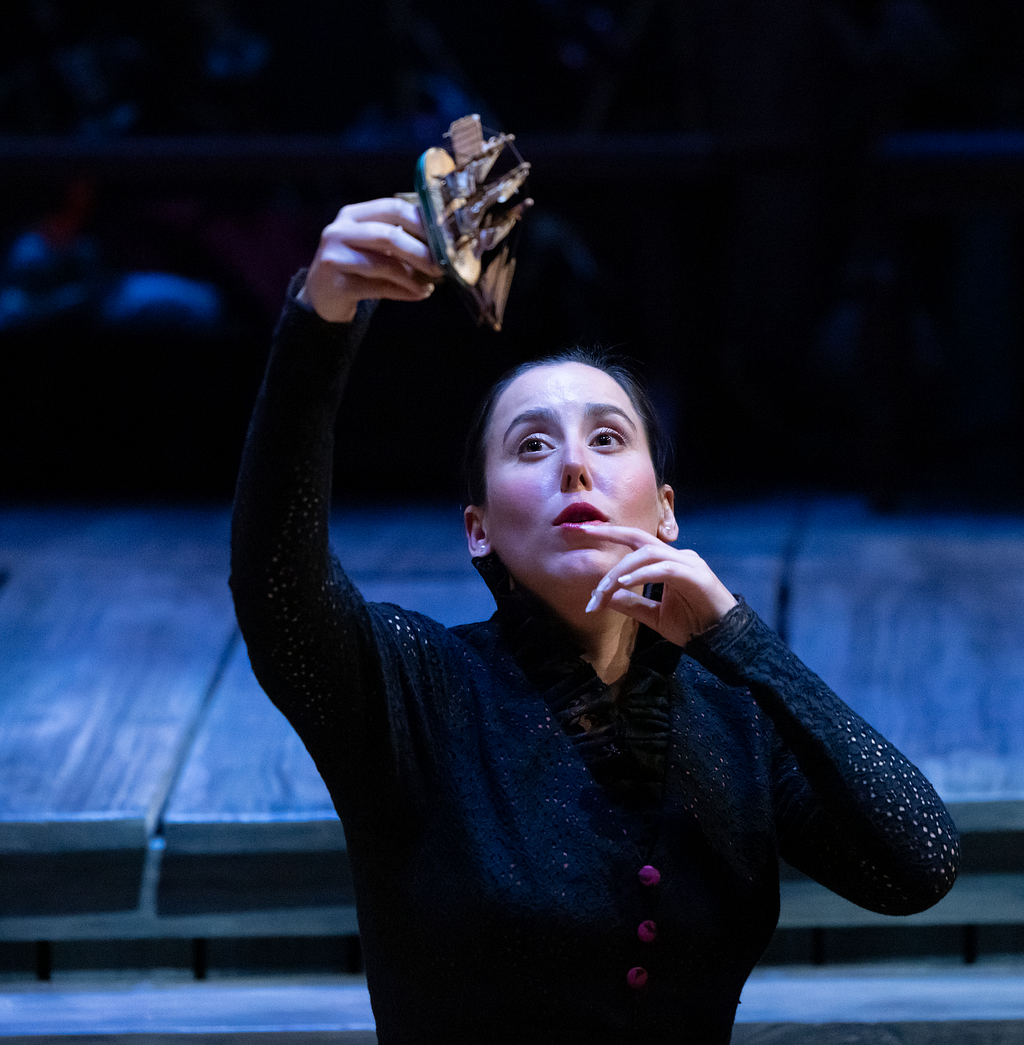 A woman in a black lacy jacket holds up a toy ship, mimicking its being tossed in a storm