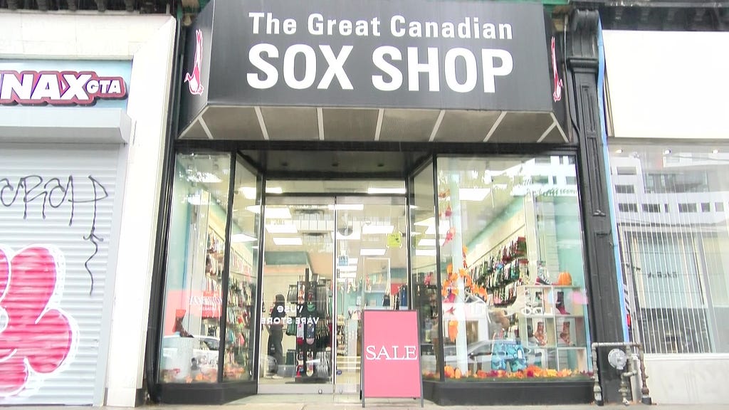 Storefront on a medium wide shot, with the store name ‘The Great Canaidan Sox Shop’. The store is open with the lights on and colourful displays seen through the window. A sign reading ‘SALE’ is placed just in front of the door.