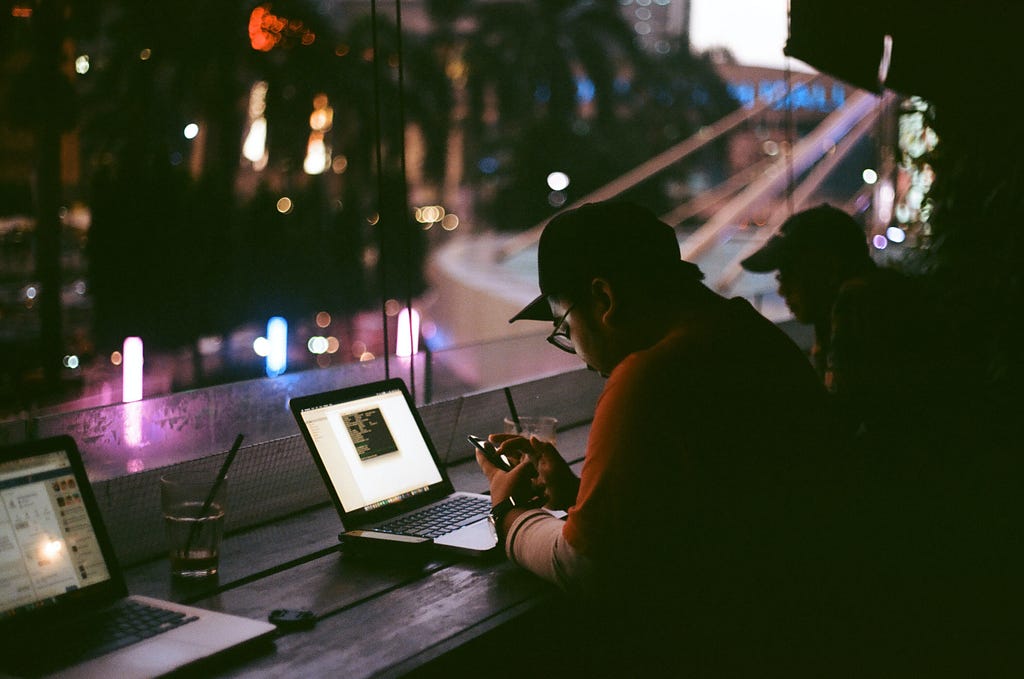 Young guys sitting at a counter with laptops looking out window at city in evening