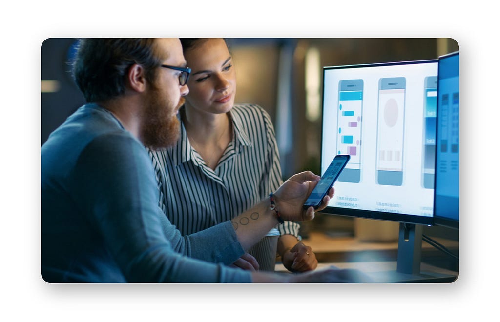 Two designers looking at mobile UI mockups on both a cell phone and a desktop, seemingly in an office.