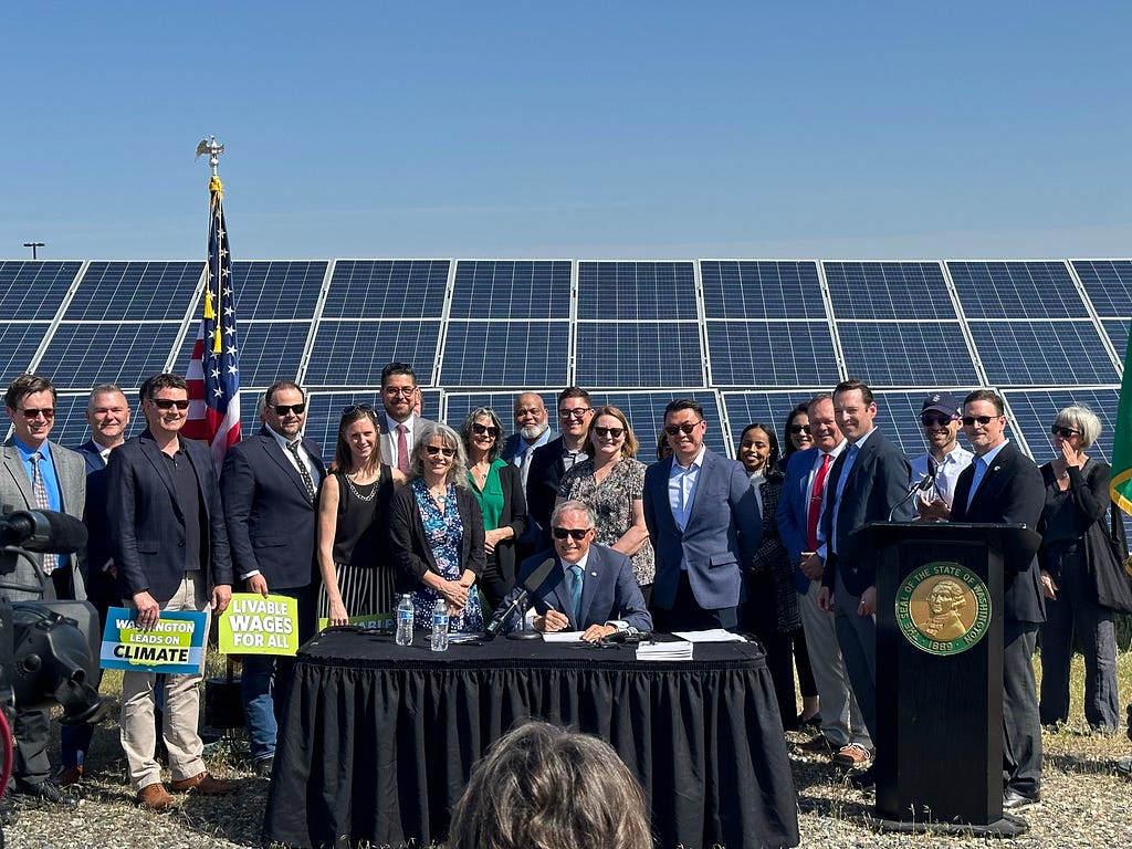 Photo of Gov. Inslee seated at a table signing a bill and a crowd of a couple dozen people standing behind him, smiling at the camera. Behind them are a large array of solar panels and bright blue sky.