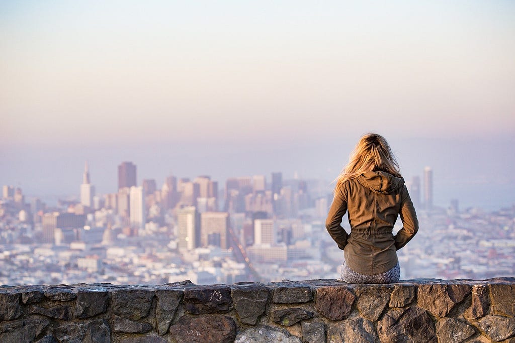 A woman sitting on a ledge, overlooking a big city.