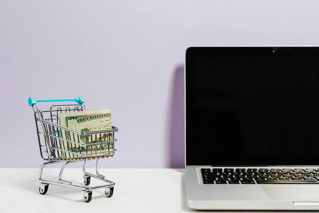 A mini doll-sized shopping cart with $20 inside the cart. The cart is sitting next to a laptop.