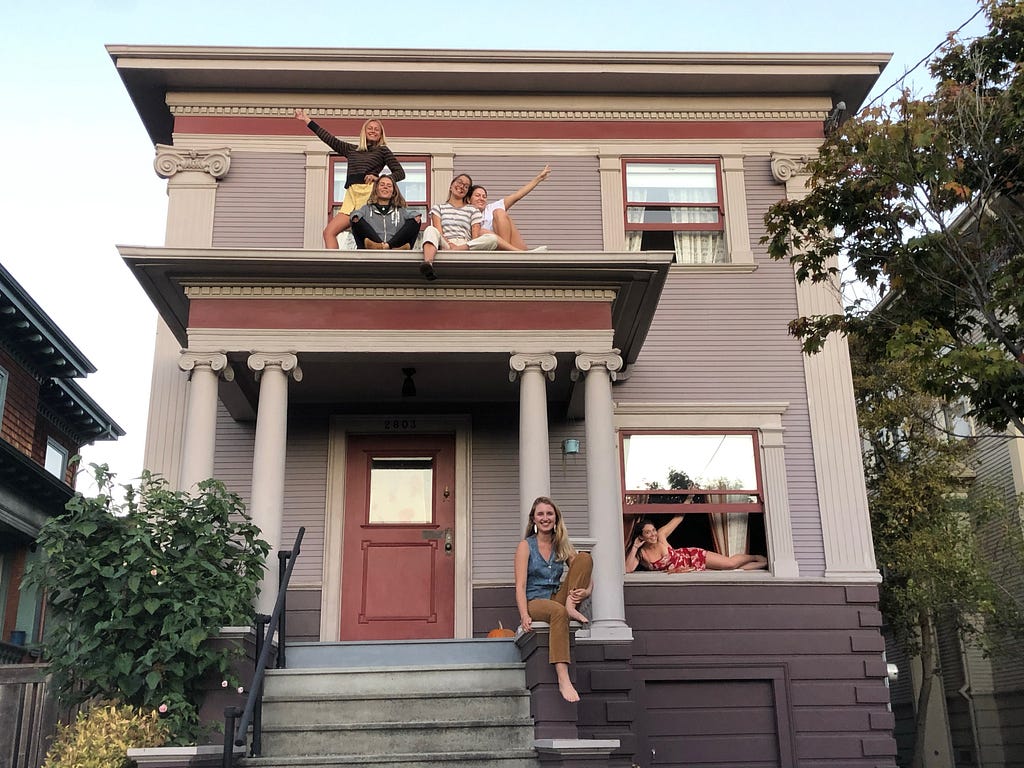 Five girls pose on and around a small purple house.