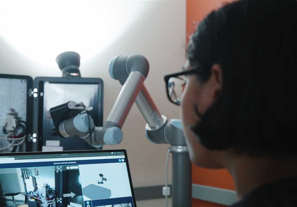 A person is using the Periscope interface in the foreground. A robotic arm with a camera views a control box in the background.