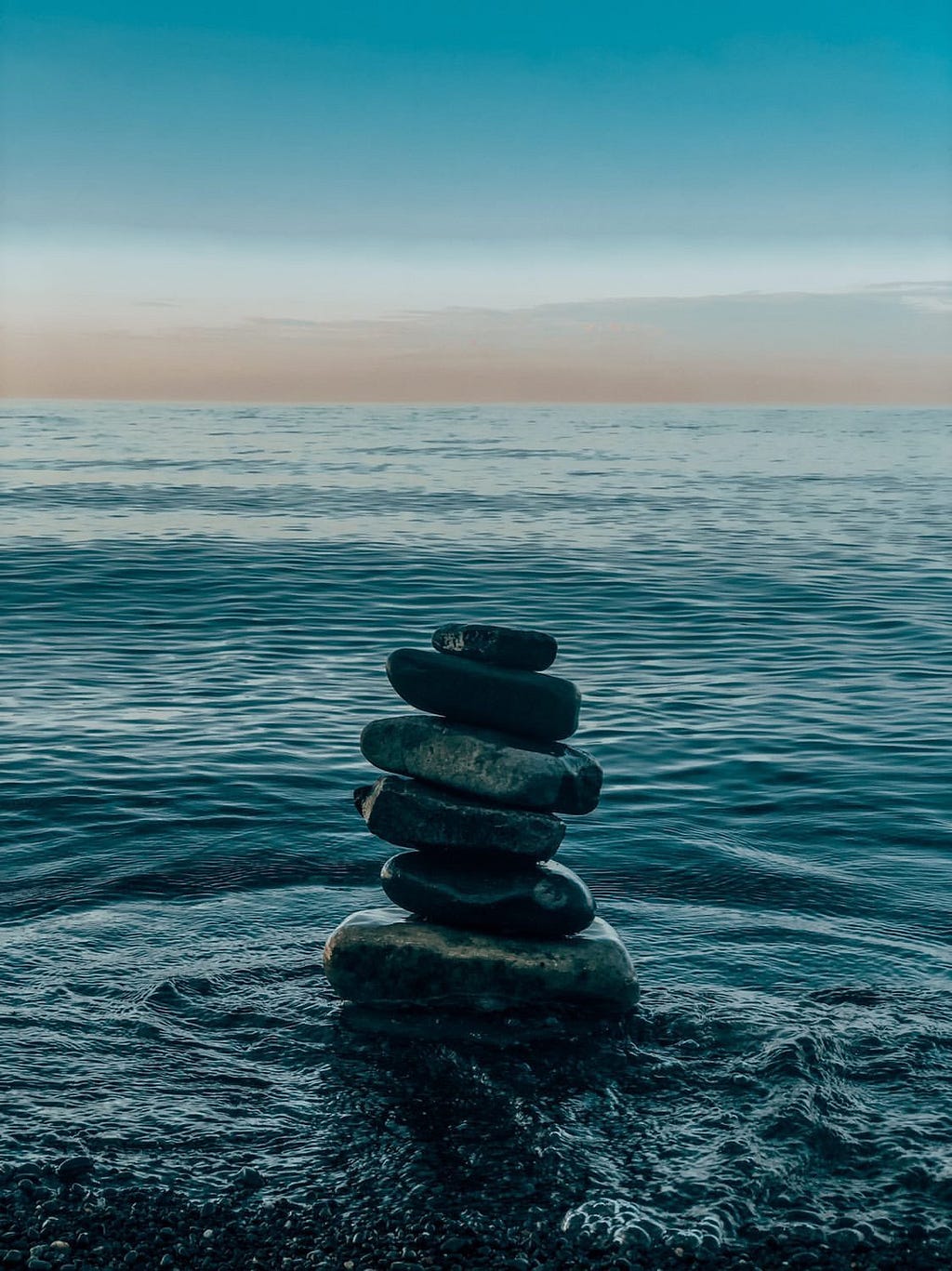 Stones stacked on top of each other surrounded by water rippling away from it. A pink horizon can be seen in the distance.