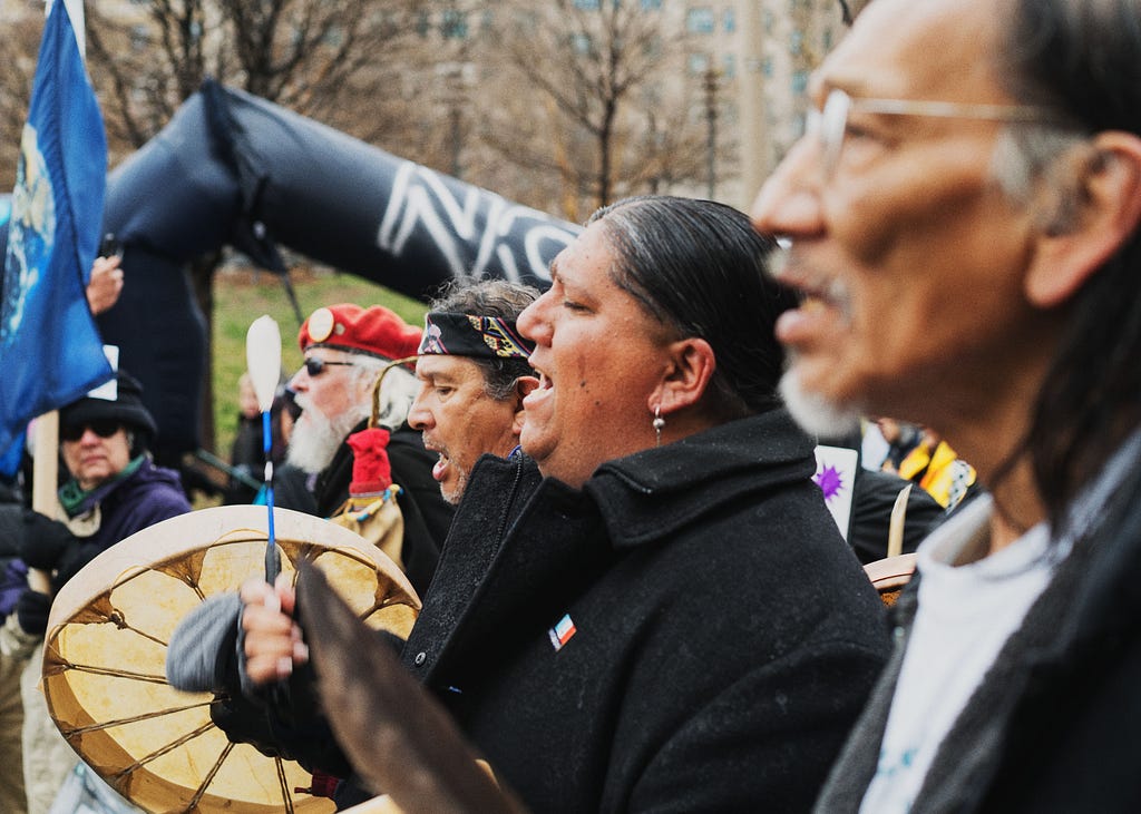 An image of four people standing in a row, taken from profile. Two people are beating drums.