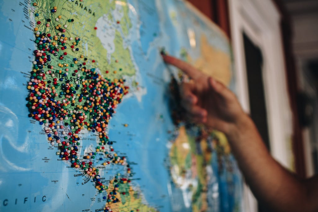 A hand pointing at a wall-mounted map of the world with numerous pushpins marking various locations.