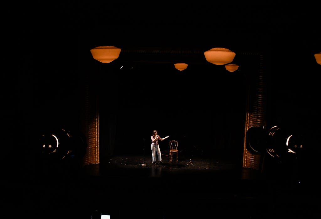 Two stage lights shine on a woman onstage. She is gesturing while performing her solo show.