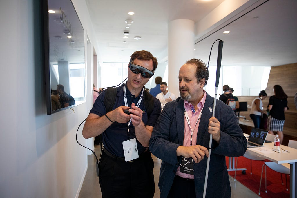 A visually impaired man wearing a Magic Leap AR device examines a controller, while another man looks on holding his white cane.