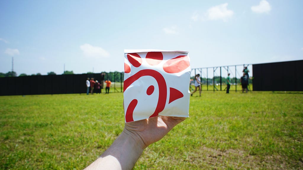 A person holding a white and red Chick-fil-a bag with a park in the background.