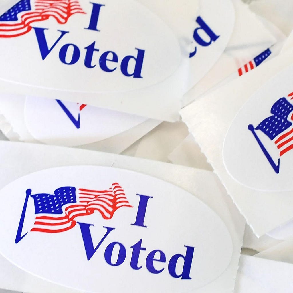 A pile of American “I Voted” stickers against a white backdrop. Each individual sticker is white with blue lettering and depicts a small artistic image of an American flag waving in the wind.