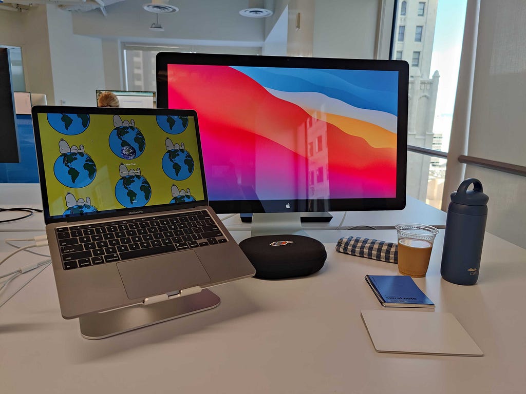 a desk with various items on it: a laptop plugged into a monitor, headphones, coffee, water bottle, and mousepad