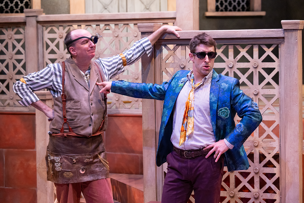 A man dressed as a jeweler leans on a staircase while another in sunglasses and a sparkly blue jacket points and looks concerned.