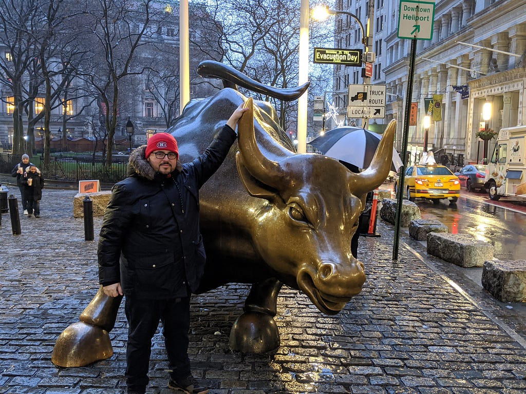 The writer of the blog, Steven Weir, stands holding the horn of a large, bronze sculpture of a charging bull near Wall Street, New York City.