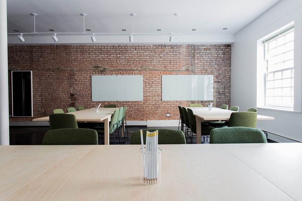 Empty office with three tables and multiple green chairs in a bright environment with a brick wall.