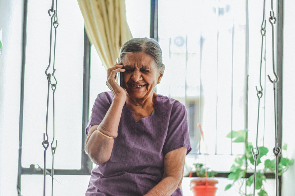 A smiling old lady sitting next to the window and having a phone call.