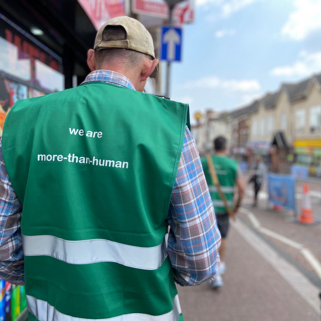 Photo of the back of a white man wearing a dark green work jacket with the words “we are more-than-human’ written on it. The man is on the pavement of Dudley High Street, there are shops in the background.