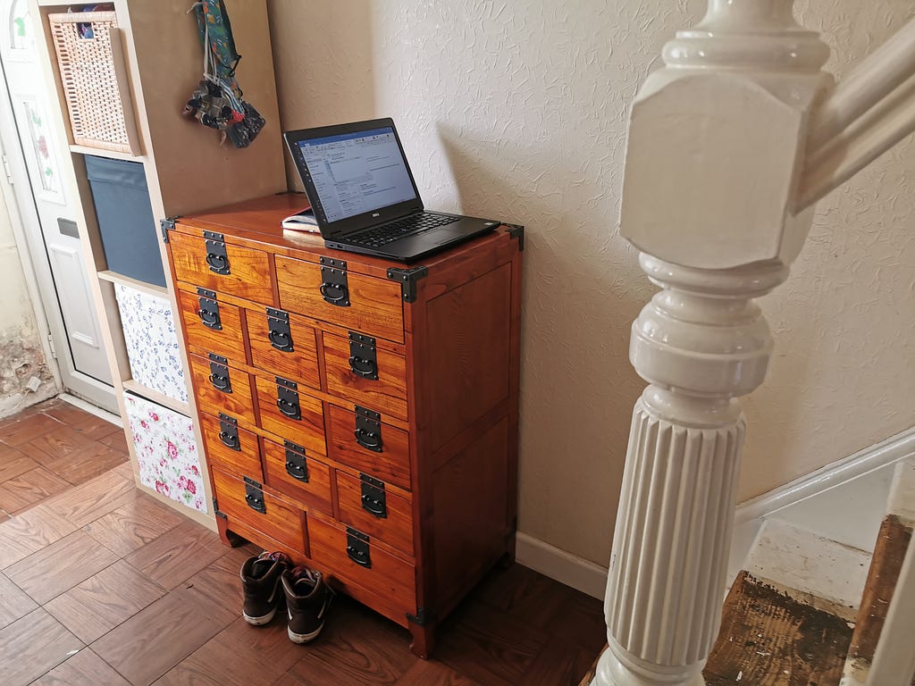 A laptop sits on the top of a vintage chest of apothecary drawers in the hallway of a house. Visible are the bottom two stairs and banister in the foreground, and in the background a taller storage unit with flowery drawers and a grubby white front door.