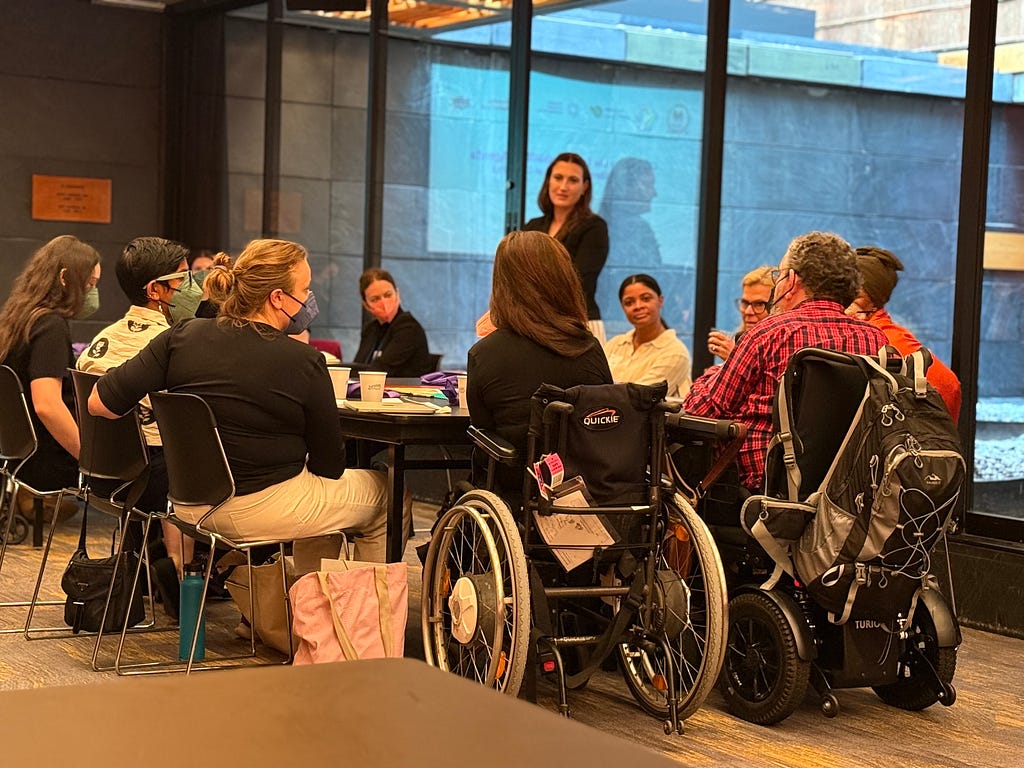 People are seated, two in wheelchairs, at a large table listening to a speaker. Many listeners wear colorful face masks, and Anastasia , WEI’s Director of Movement and Capacity Strengthening, stands in the background.