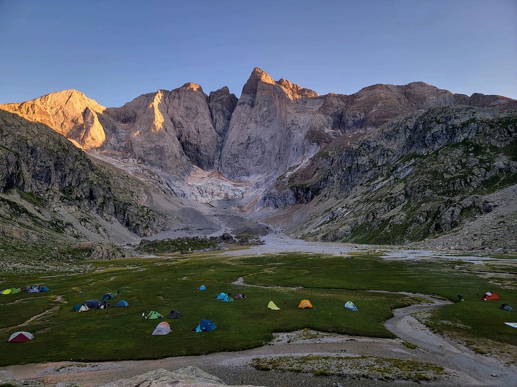 A group of tents on a flat plain underneath a large stone mountain. The sun is reflecting of its upper left peak. Taken at Vignemale in the Parc National des Pyrénées.