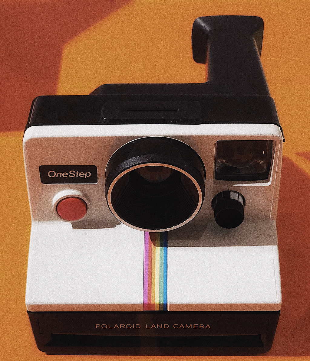 White Polaroid Land Camera with a One Step red button and rainbow running down the middle. This image has an orange background.