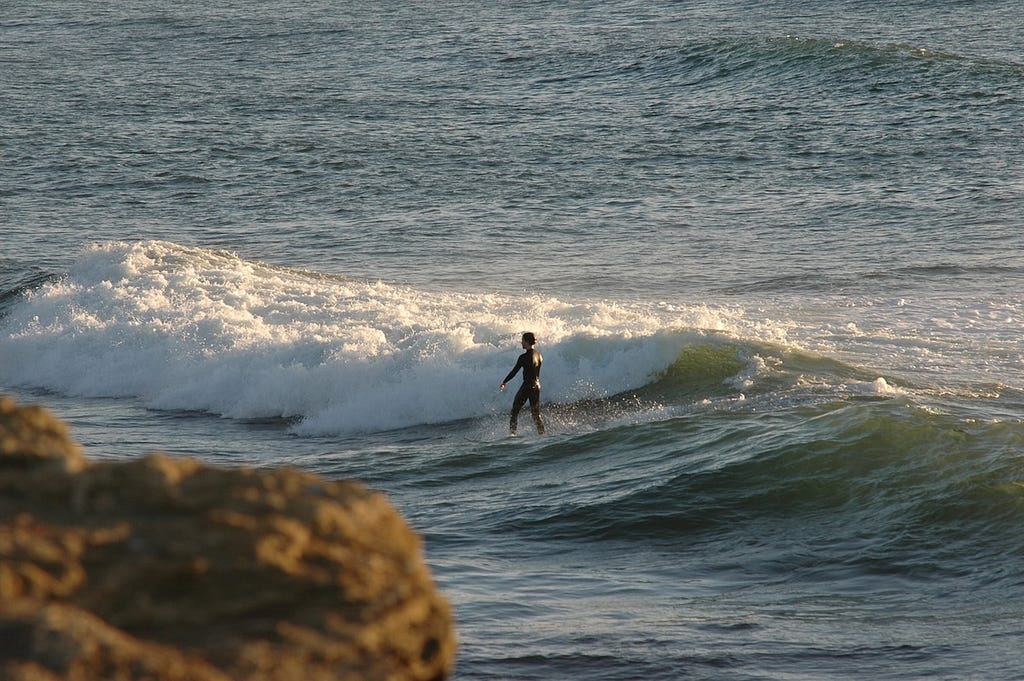A man surfing an ocean wave in the middle of the picture, with a blurry rocky outcrop in the foreground on the left.